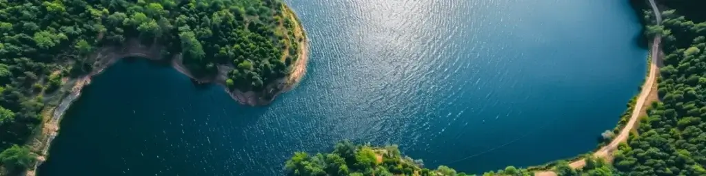 Flygfoto över en slingrande sjö omgiven av täta, grönskande skogar. Vattnet är djupblått, med solljus som reflekteras från dess yta. Strandlinjer är markerade av klippor och grönska, vilket skapar ett fridfullt och pittoreskt landskap.