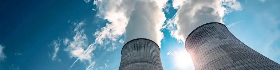 Bilden visar två stora kyltorn som avger ånga mot en klarblå himmel med några spridda moln. Tornen är en del av en industrianläggning, troligen ett kraftverk, med solljus som lyser upp ångan.