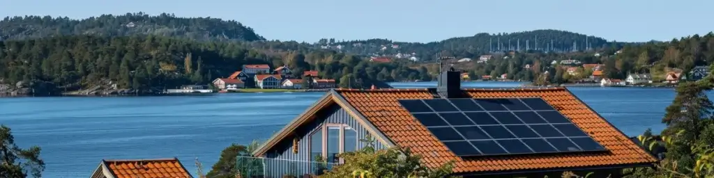 En naturskön sjöutsikt med hus utspridda längs strandlinjen och kullar i bakgrunden. I förgrunden syns ett hus med rött tegeltak och Monokristallina solceller väl synligt, omgivet av grönska, med den lugna sjön och skogsklädda kullar bakom sig.