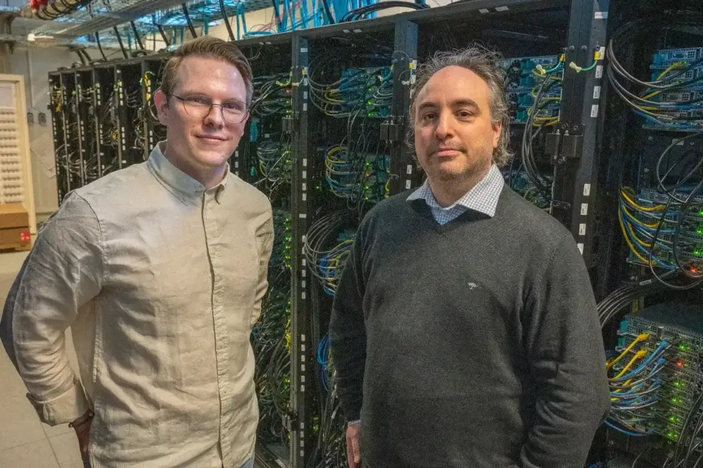 Två män står framför en rad datorservrar i ett datacenter. Mannen till vänster bär glasögon och en ljus skjorta med knäppning, medan mannen till höger bär en mörk tröja över en krage. Båda är vända mot kameran.